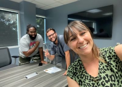 Three people smiling in a meeting room; one taking a selfie. A table and TV are in the background.