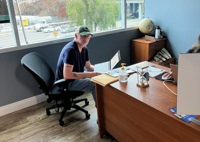 Person in a cap sitting at a desk with a laptop, looking up. Office with a window view of a road outside.