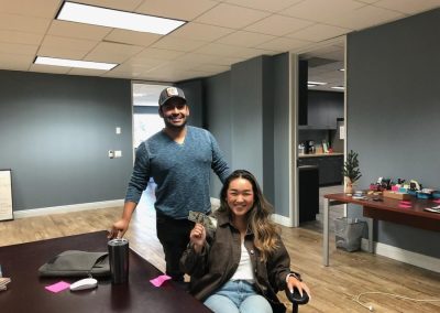 Smiling woman holding money sits in an office chair; a man stands beside her, both in a well-lit office space.