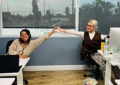 Two people sitting at desks, smiling and making a heart shape with their hands in an office with large windows.