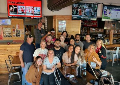 A group of people smiling and posing inside a bar with sports on TV screens above them.