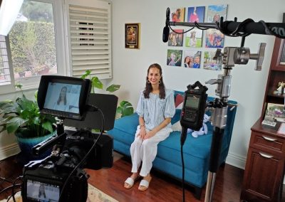 Woman sitting on a blue sofa in a home studio setup with cameras and lights. Photos on the wall in the background.