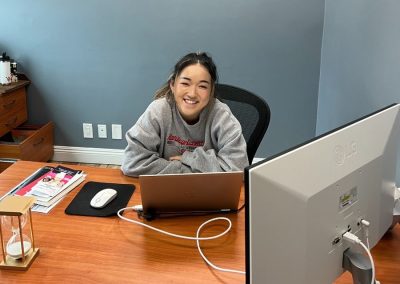 Person sitting at a desk with a laptop and monitor, smiling at the camera in an office setting.