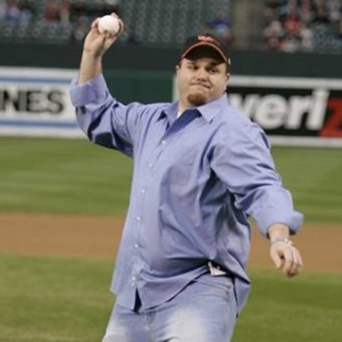 jeremy_01 Man in a blue shirt and cap throwing a baseball on a field, with stands in the background.