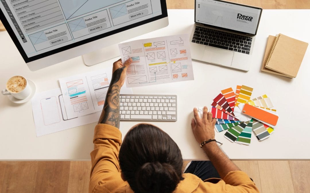 Person designing a website at a desk with color swatches, sketches, and a computer displaying web layouts.