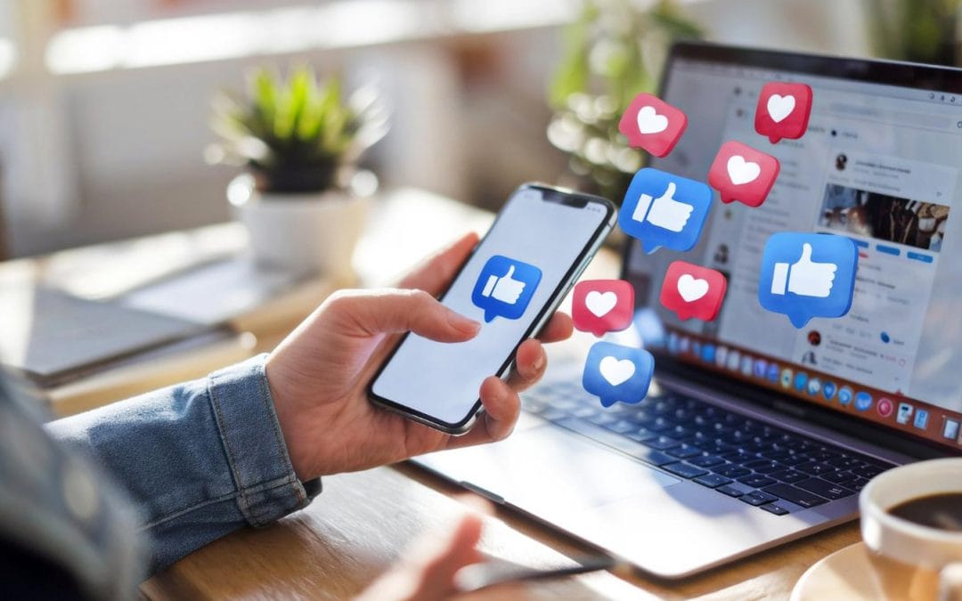 Person holding smartphone next to laptop with social media like and heart icons floating in the air.