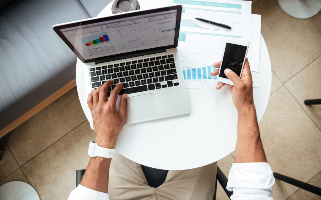 Person working at a laptop while holding a smartphone, with charts and a coffee cup on a white table.